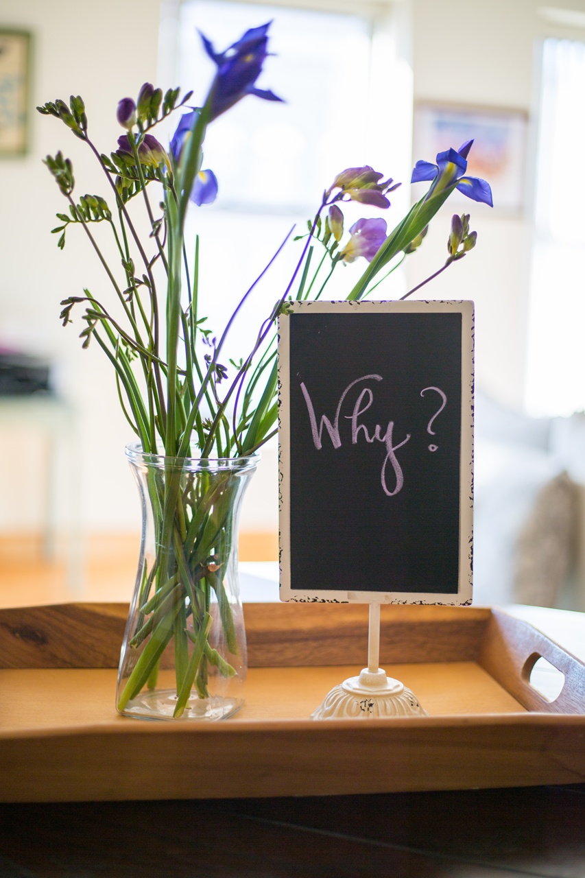 Schild mit der Schrift “Why?” auf einem Tablett neben einem Strauß Blumen in einer Vase