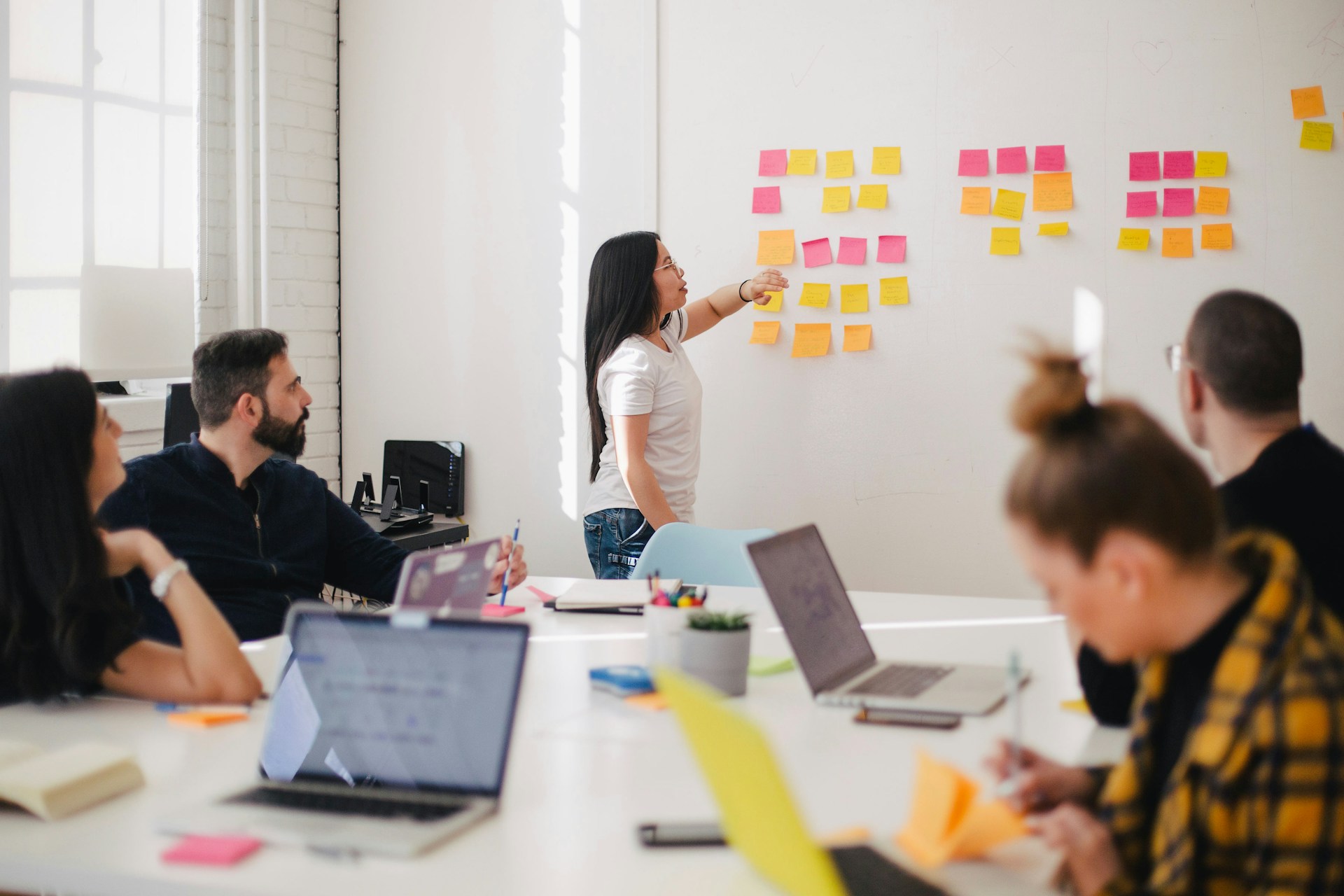 Frau moderiert ein Team-Meeting stehend vor einer Wand mit angeklebten bunten Klebezetteln"